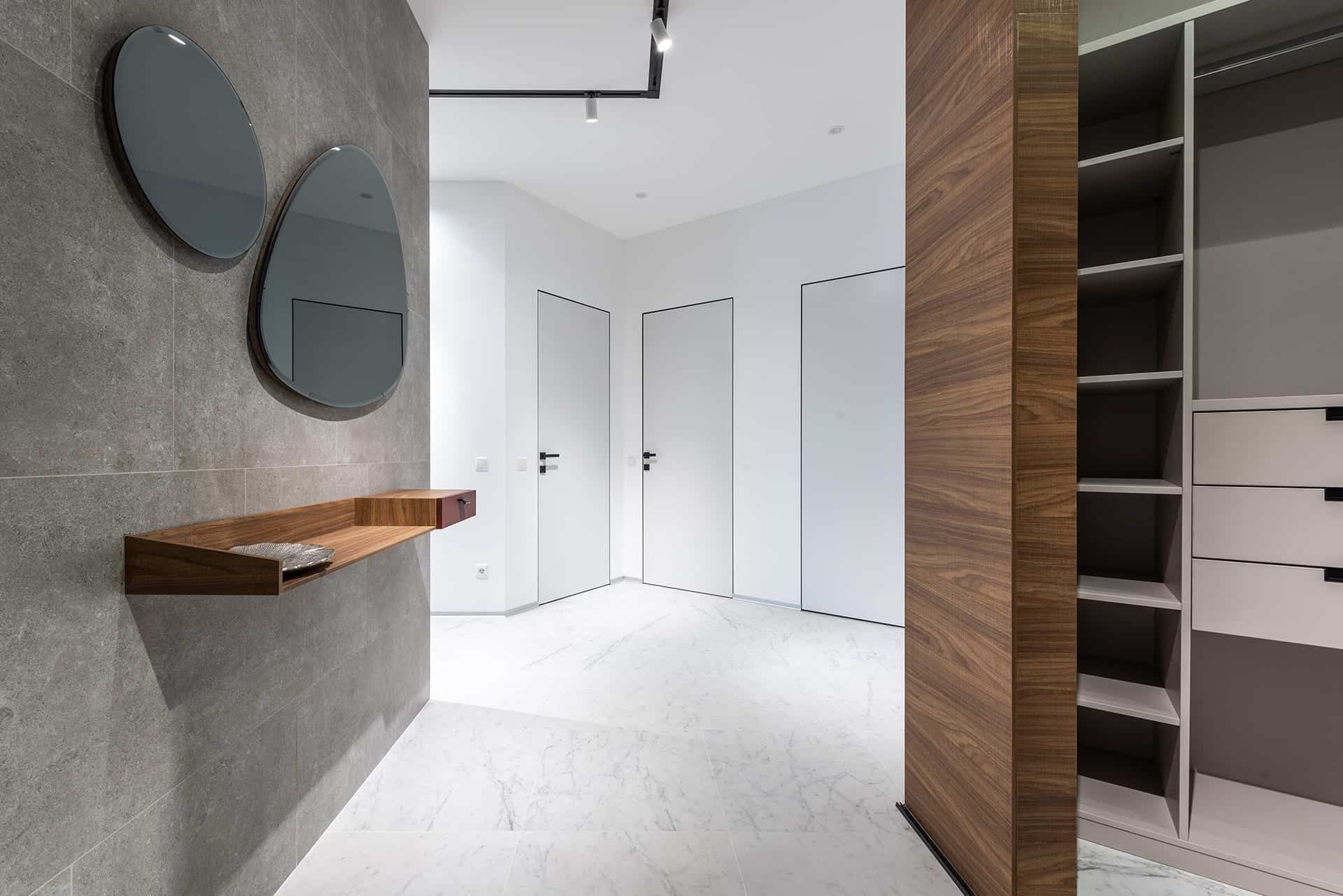Modernes Foyer-Design mit grauer Wand in Betonoptik, organisch geformten Spiegeln, schwebendem Holzregal, weißem Marmorboden und einem geöffneten Einbauschrank mit Holzverkleidung.