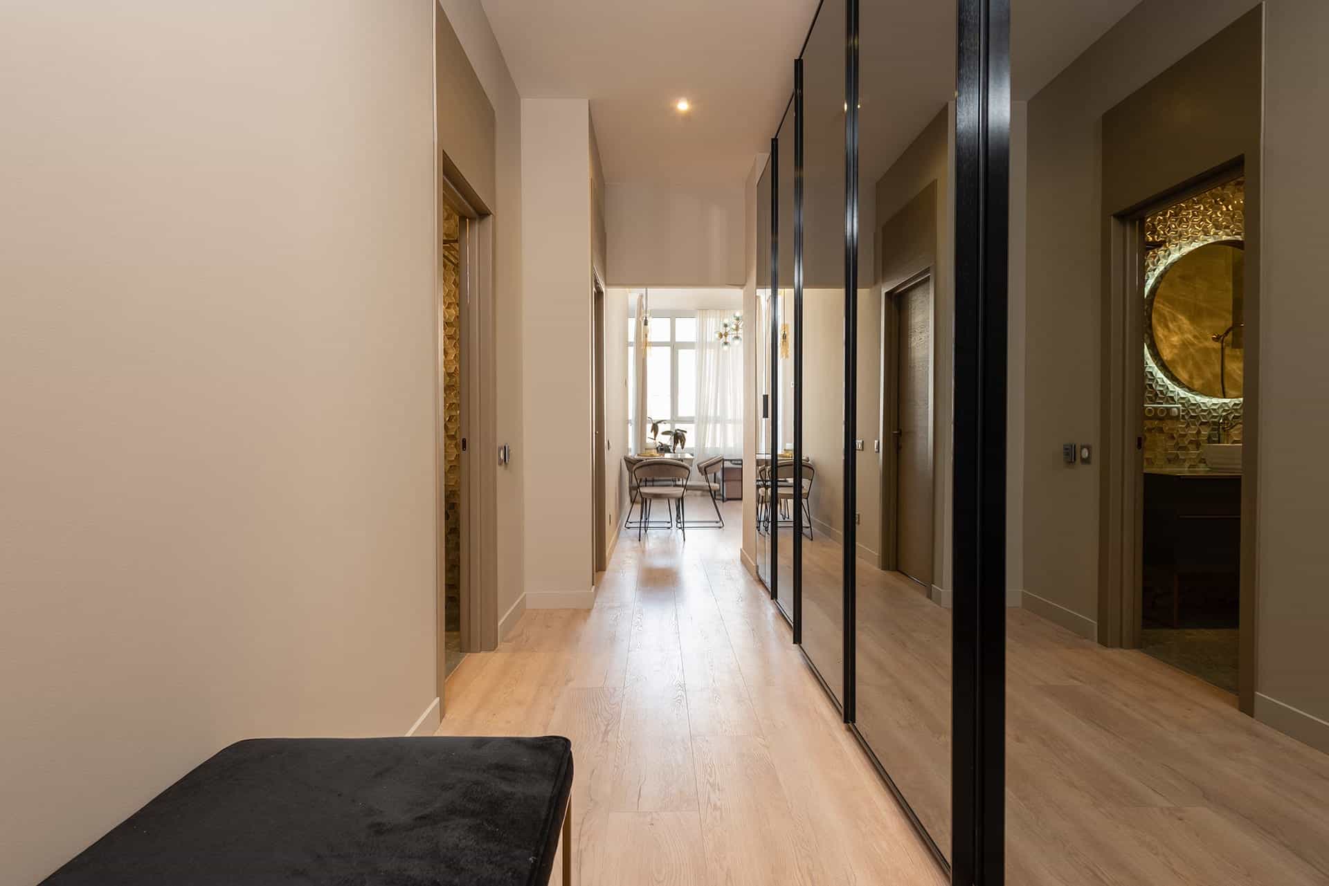 Langer moderner Flur mit hellem Holzboden, großem Spiegelschrank mit schwarzem Rahmen, beigefarbenen Wänden und Blick auf ein helles Esszimmer.