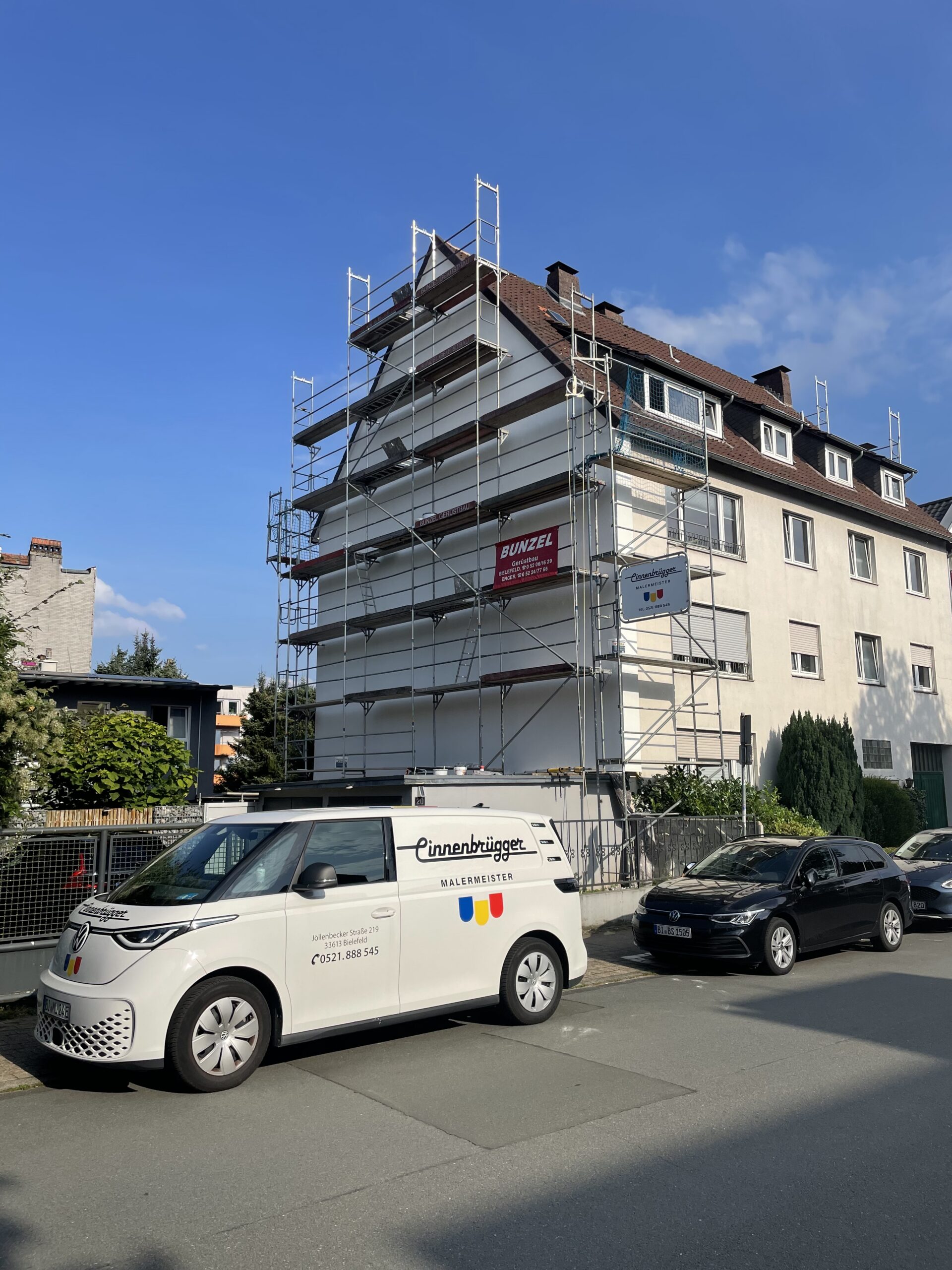 Weißer VW ID. Buzz Firmenwagen von Linnenbrügger Malermeister vor einem eingerüsteten Mehrfamilienhaus bei der Fassadensanierung unter blauem Himmel.
