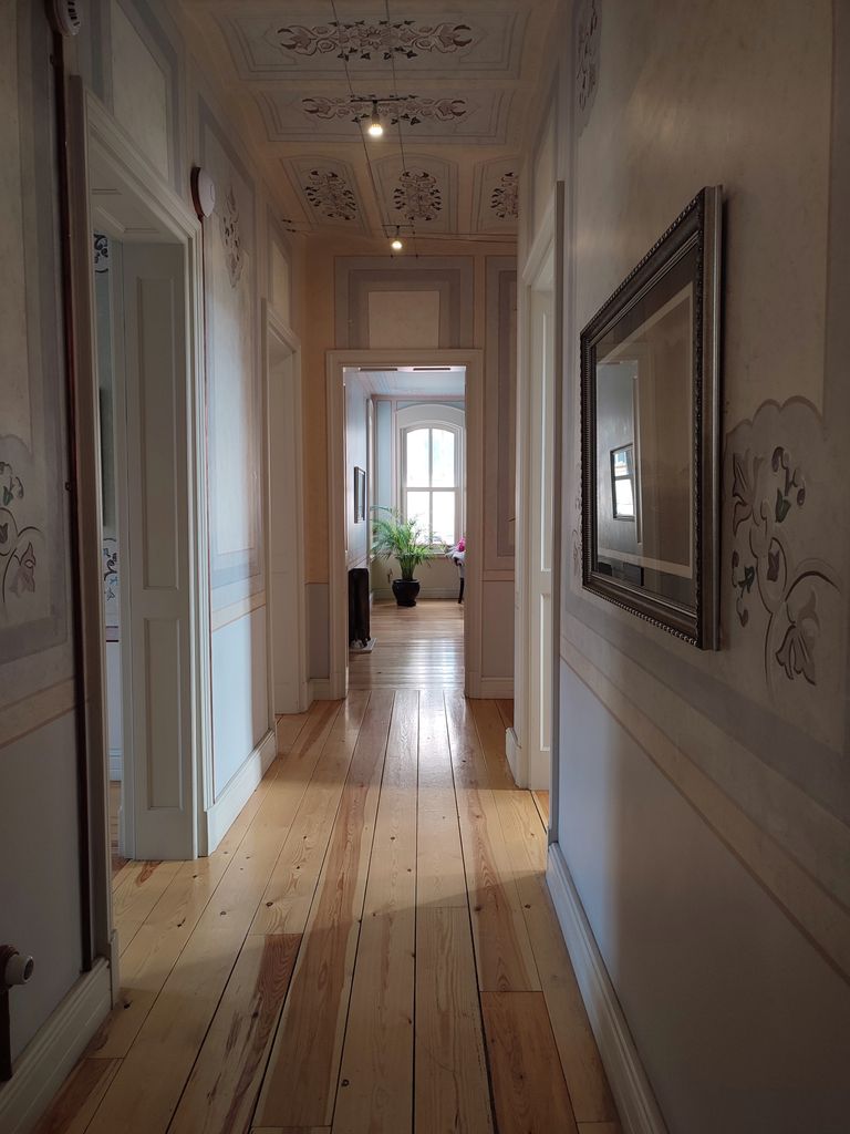 Langer, historischer Flur mit echten Holzdielen, kunstvoller Deckenmalerei, floralen Wandornamenten und Blick in ein angrenzendes Zimmer.