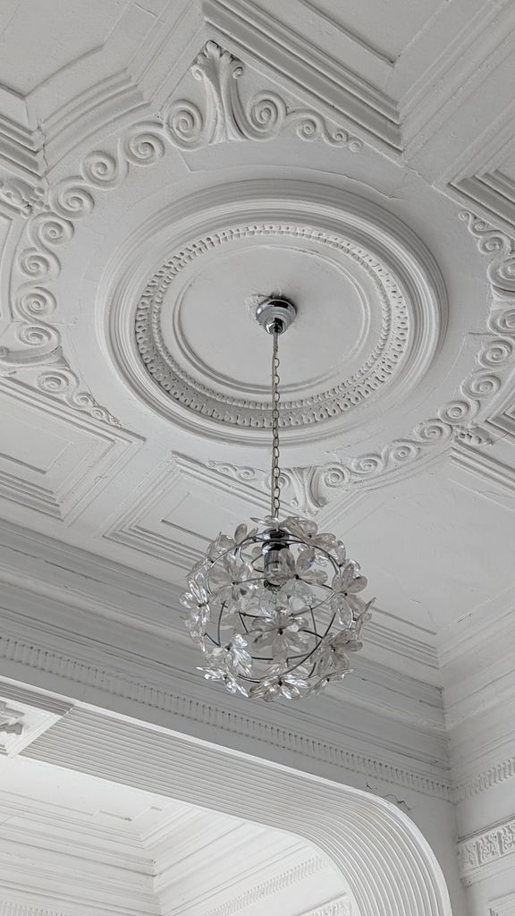 Nahaufnahme einer hochweißen Altbau-Stuckdecke mit runder Deckenrosette, feinen Ornamenten und einem herabhängenden Kristall-Kronleuchter.