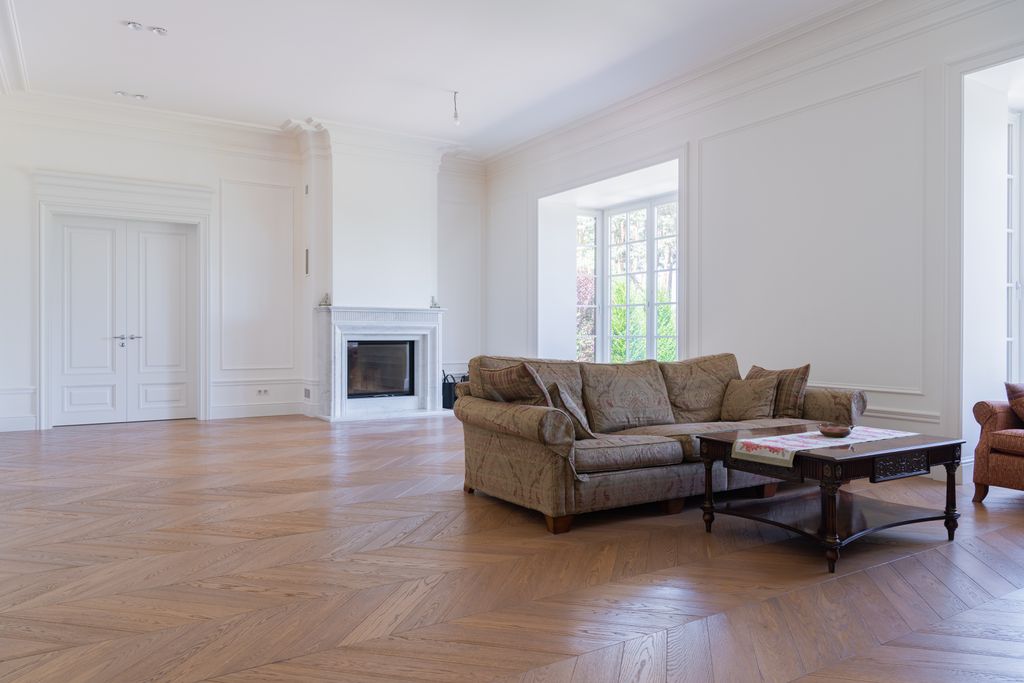 Weitläufiges, weißes Wohnzimmer mit französischem Fischgrät-Parkett, einem gemauerten Kamin, weißen Flügeltüren und einem beigen Sofa mit Couchtisch.