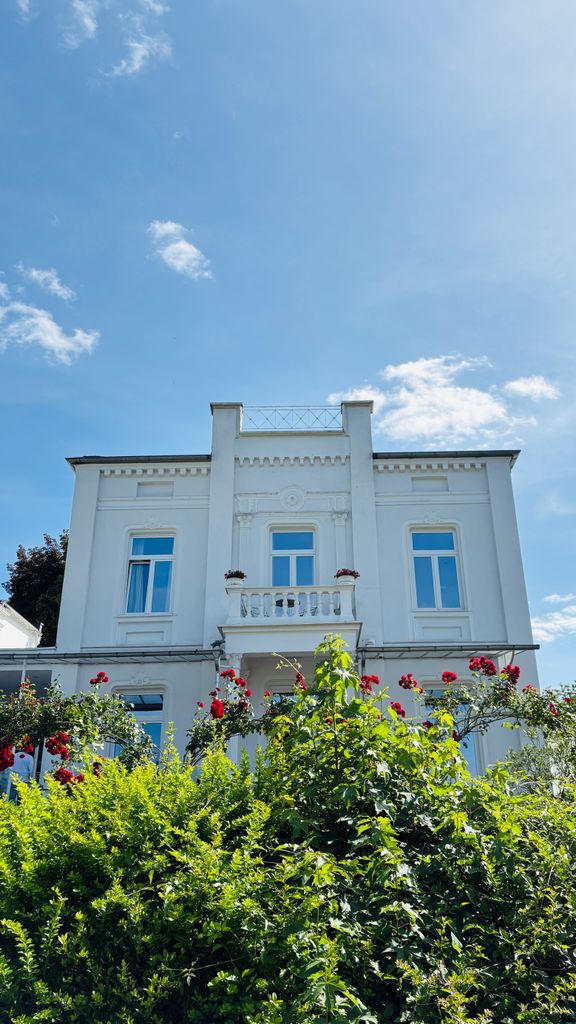 Symmetrische Außenansicht einer weiße, historischen Villa mit Balkon, Balustrade, aufwendigen Friesen und roten Rosen im Vordergrund vor blauem Himmel.