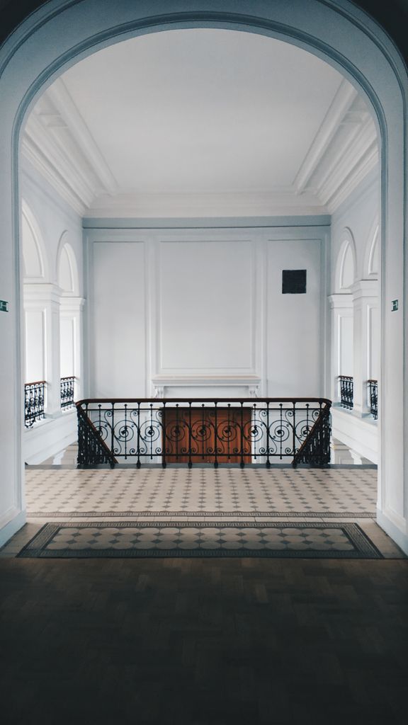 Symmetrischer Blick in ein historisches Treppenhaus mit filigranem schmiedeeisernen Geländer, weiße Wandkassetten, Säulenbögen und Fliesenboden.