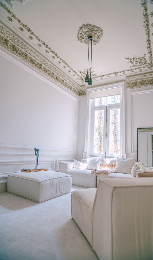 Lichtdurchflutetes, weißes Wohnzimmer mit aufwendigen Wandstuck-Profilen, einem beigen Polstersofa mit Kissen und einem großen Sprossenfenster.